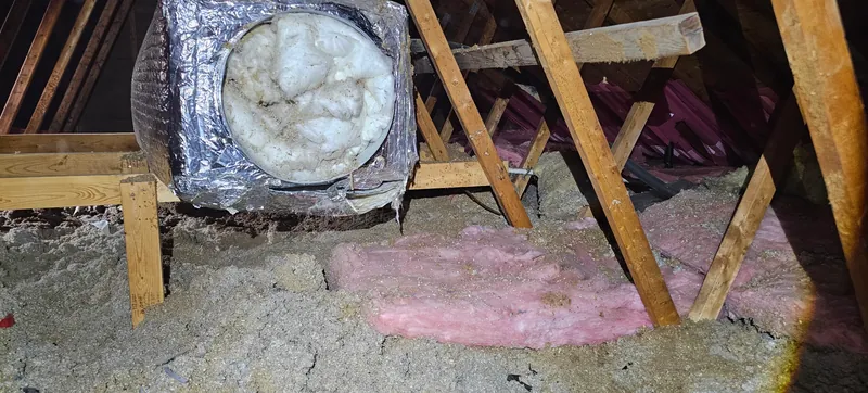 Grenier avant les travaux : ancien isolant en vrac gris-beige, avec des restes de nattes roses, autour d'un conduit enveloppé d'aluminium posé sur un support de bois