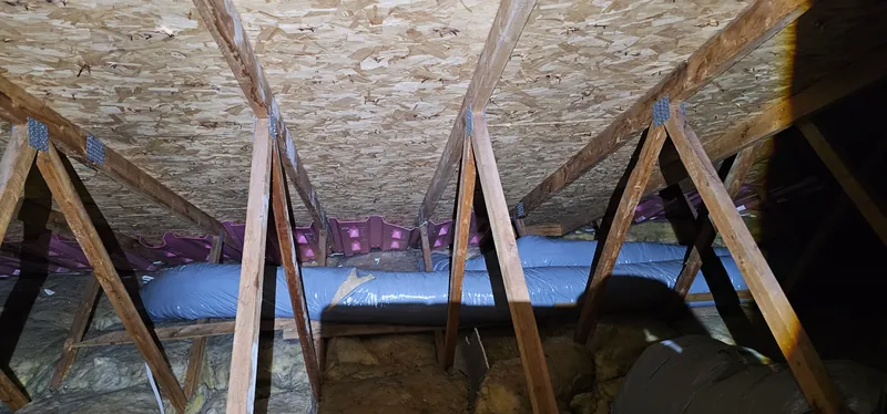 Grenier avant les travaux : vieilles nattes de laine jaune, tassées au pied des fermes de toit, sous un long conduit souple gris, déflecteurs roses au bord du toit