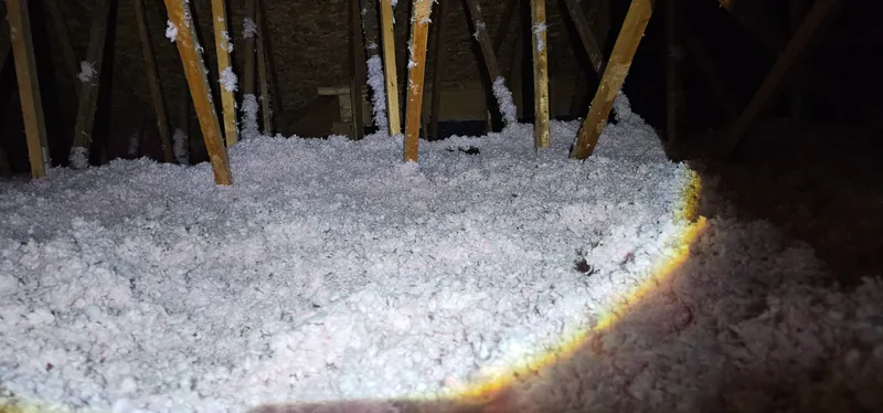 Grenier après les travaux : fibre de verre soufflée rosée, épaisse et uniforme, entre les montants des fermes, éclairée à la lampe de poche