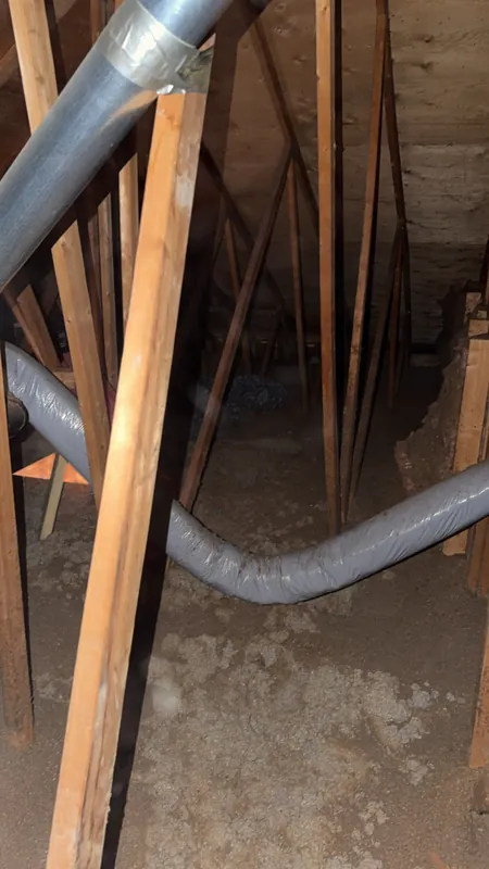 Grenier avant les travaux : ancien isolant en vrac gris-beige, tassé, sur lequel traîne un conduit souple gris, entre les fermes de toit