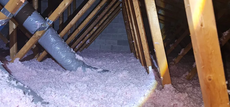 Grenier après les travaux : fibre de verre soufflée rose, épaisse, qui enfouit à moitié les mêmes conduits souples, mur de blocs de béton au fond