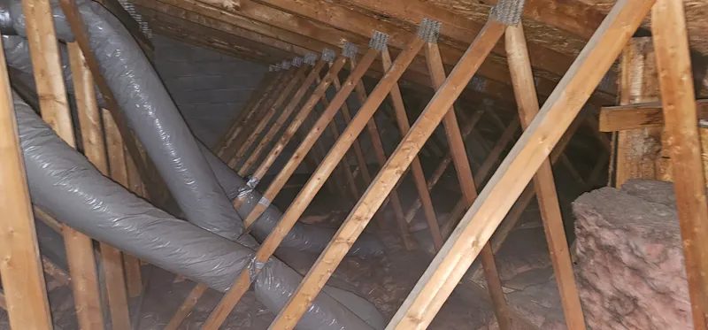 Grenier avant les travaux : ancien isolant gris et poussiéreux sous des conduits souples gris accrochés aux fermes de toit, mur de blocs de béton au fond